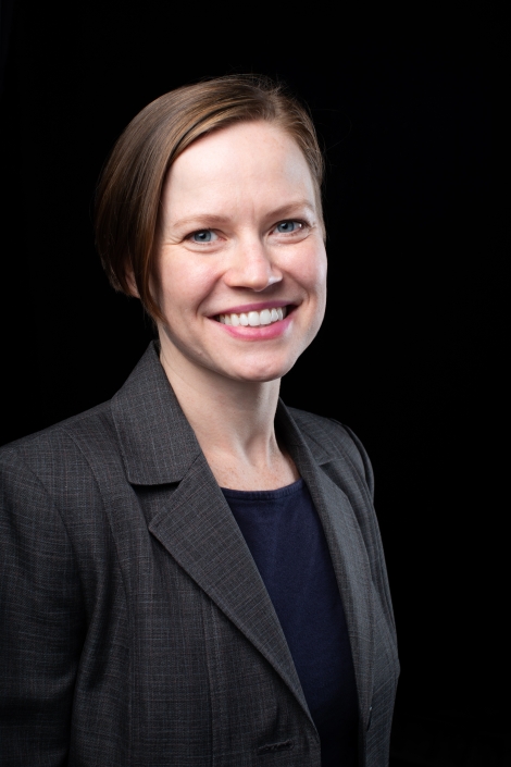 Portrait of Leah Hamilton. The image is a close up headshot with a black background. Leah has short brown hair, is smiling, and wearing a navy shirt with a gray blazer over it.